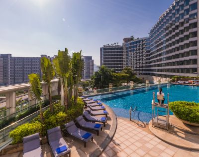 Outdoor pool area with round umbrella-shaded loungers, lush greenery, and high-rise buildings in the background. The scene is tranquil and sunny.