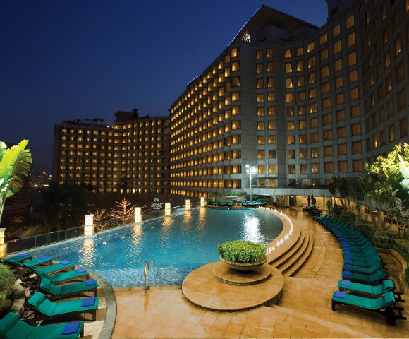 Night view of a luxurious hotel pool area. The curved pool is surrounded by green lounge chairs, and the well-lit hotel facade creates a welcoming atmosphere.