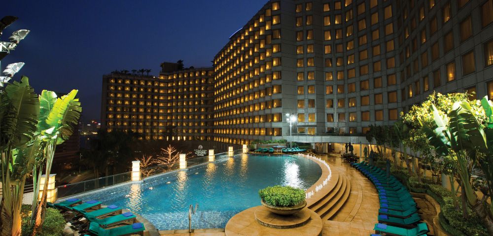 Night view of a luxurious hotel pool area. The curved pool is surrounded by green lounge chairs, and the well-lit hotel facade creates a welcoming atmosphere.