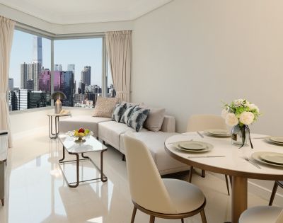 Bright, modern living room with city skyline view. Features a beige sofa, dining table set, cushioned chairs, and decorative flowers, exuding elegance.