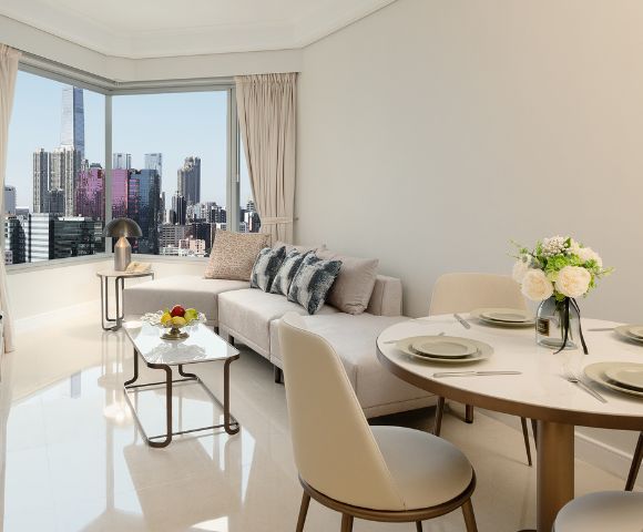 Bright, modern living room with city skyline view. Features a beige sofa, dining table set, cushioned chairs, and decorative flowers, exuding elegance.