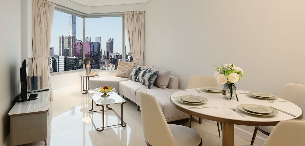 Bright, modern living room with city skyline view. Features a beige sofa, dining table set, cushioned chairs, and decorative flowers, exuding elegance.