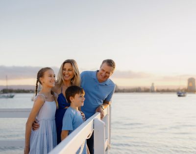 A family of four smiles on a pier during sunset. The parents and two children are dressed in blue, with calm water and distant sailboats in the background.