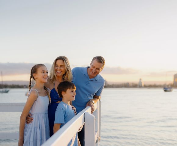 A family of four smiles on a pier during sunset. The parents and two children are dressed in blue, with calm water and distant sailboats in the background.