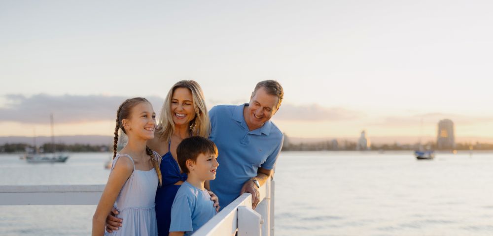 A family of four smiles on a pier during sunset. The parents and two children are dressed in blue, with calm water and distant sailboats in the background.