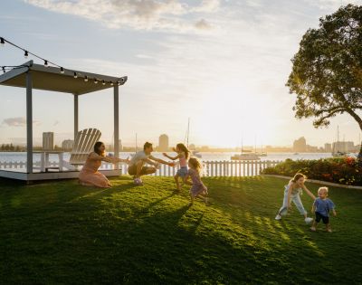 A family with children plays joyfully on a sunlit lawn near a white fence. A gazebo and string lights add charm, as a city skyline looms in the background.