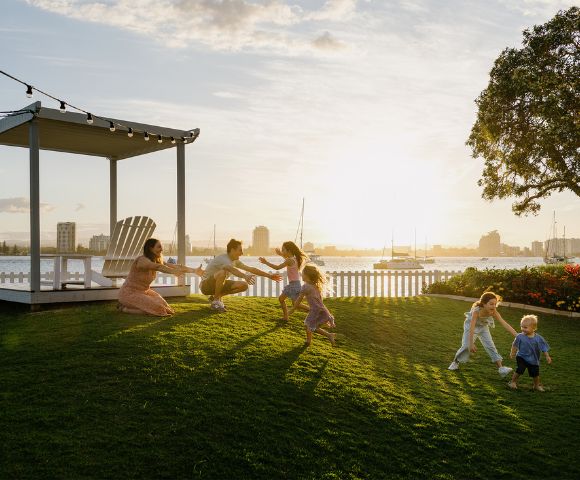 A family with children plays joyfully on a sunlit lawn near a white fence. A gazebo and string lights add charm, as a city skyline looms in the background.