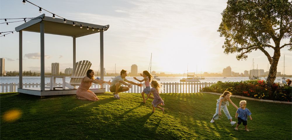 A family with children plays joyfully on a sunlit lawn near a white fence. A gazebo and string lights add charm, as a city skyline looms in the background.