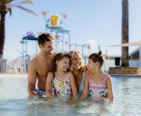 A joyful family of four, wearing swimsuits, enjoys a sunny day in a pool. Palm trees and colorful water park structures are visible in the background.