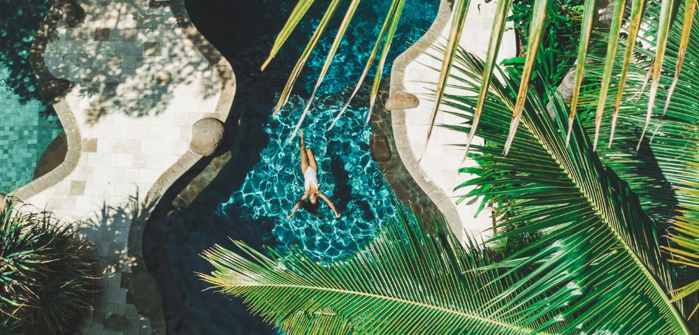 A woman swims in a pool, surrounded by lush palm trees under a clear blue sky.
