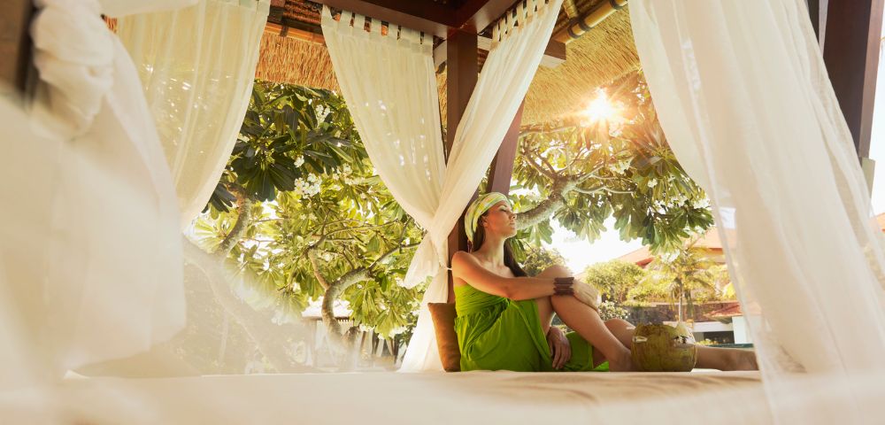 A woman sitting on a bed in a bright white room, looking relaxed and comfortable.
