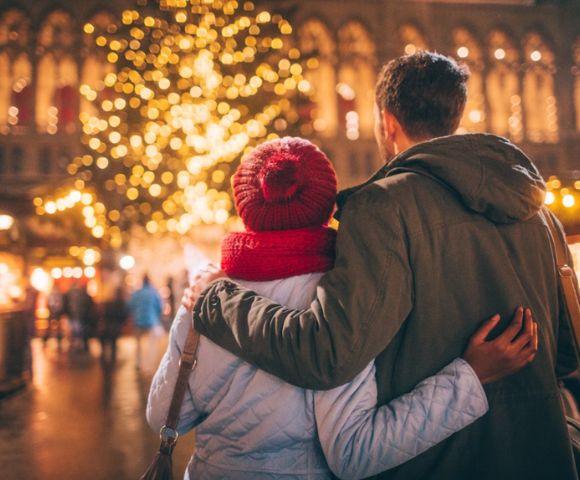 A couple, seen from behind, embraces warmly in a festive holiday market. They face a beautifully lit Christmas tree, creating a cozy, joyful atmosphere.