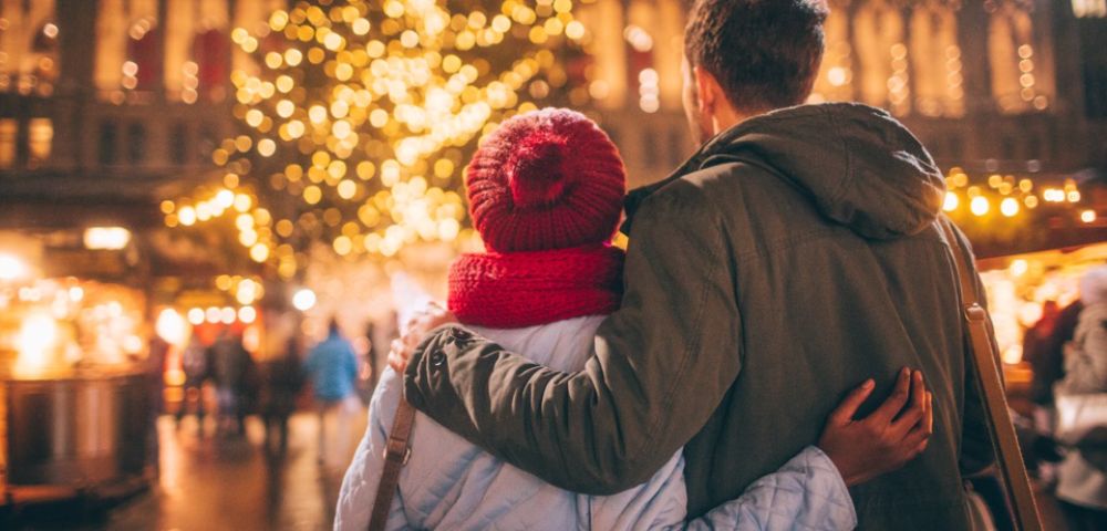 A couple, seen from behind, embraces warmly in a festive holiday market. They face a beautifully lit Christmas tree, creating a cozy, joyful atmosphere.