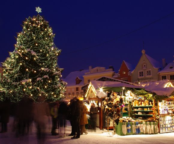 Festive Christmas market scene at night with a large illuminated tree, cozy stalls selling gifts, and blurred people creating a warm, cheerful atmosphere.