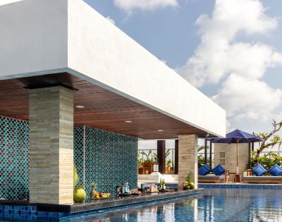 Outdoor poolside bar with turquoise mosaic tiles, stocked shelves, and bar stools. Sunny weather, blue sky, and loungers under purple umbrella. Relaxing vibe.