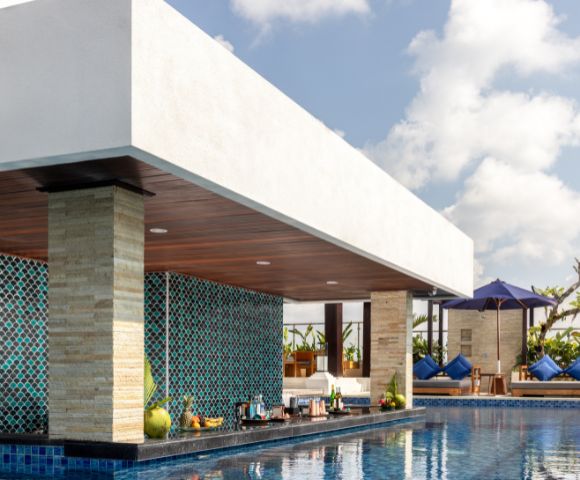 Outdoor poolside bar with turquoise mosaic tiles, stocked shelves, and bar stools. Sunny weather, blue sky, and loungers under purple umbrella. Relaxing vibe.