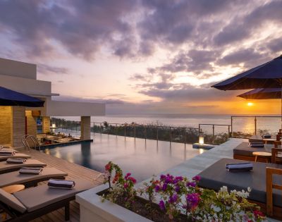 Sunset view from a luxurious rooftop pool deck, featuring lounge chairs, vibrant flowers, and umbrellas. The sky is a blend of orange, purple, and pink hues.