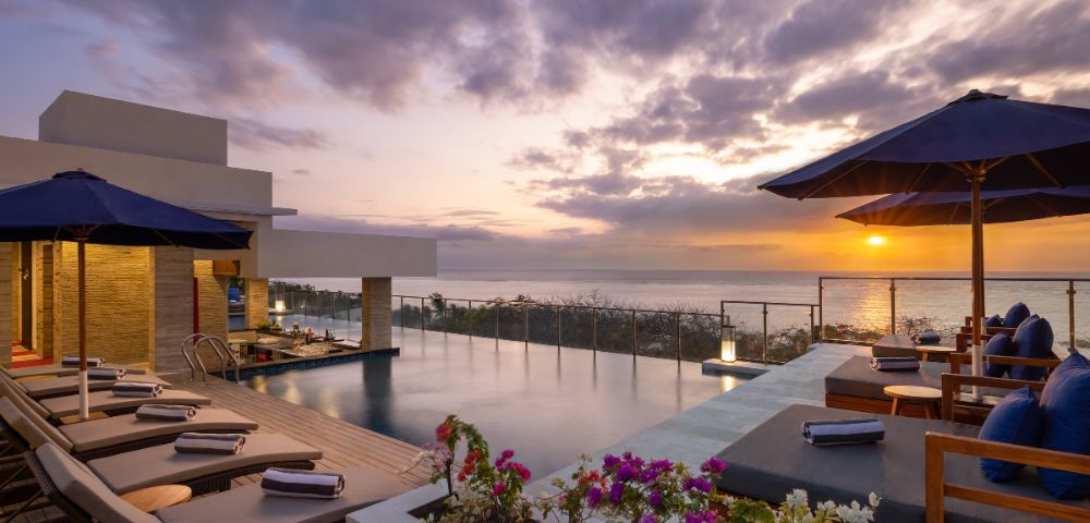 Sunset view from a luxurious rooftop pool deck, featuring lounge chairs, vibrant flowers, and umbrellas. The sky is a blend of orange, purple, and pink hues.