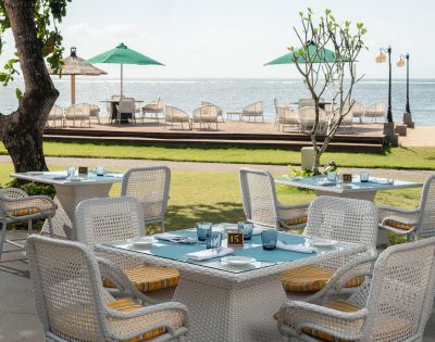 Outdoor dining area with wicker tables and chairs facing the ocean. Green umbrellas and deck chairs line the wooden deck. A tranquil, sunny atmosphere.