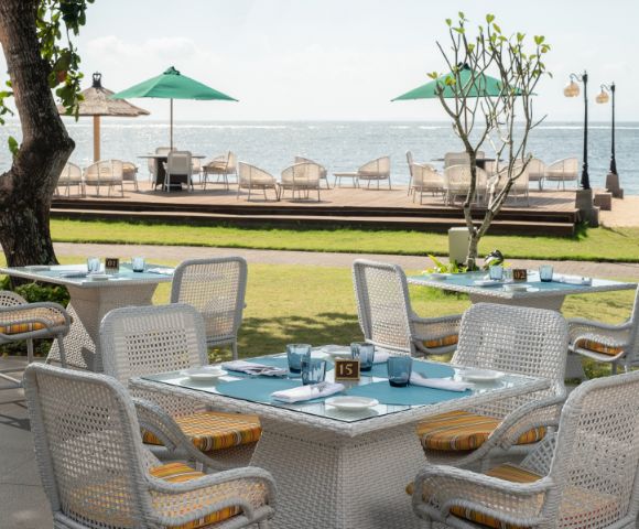 Outdoor dining area with wicker tables and chairs facing the ocean. Green umbrellas and deck chairs line the wooden deck. A tranquil, sunny atmosphere.
