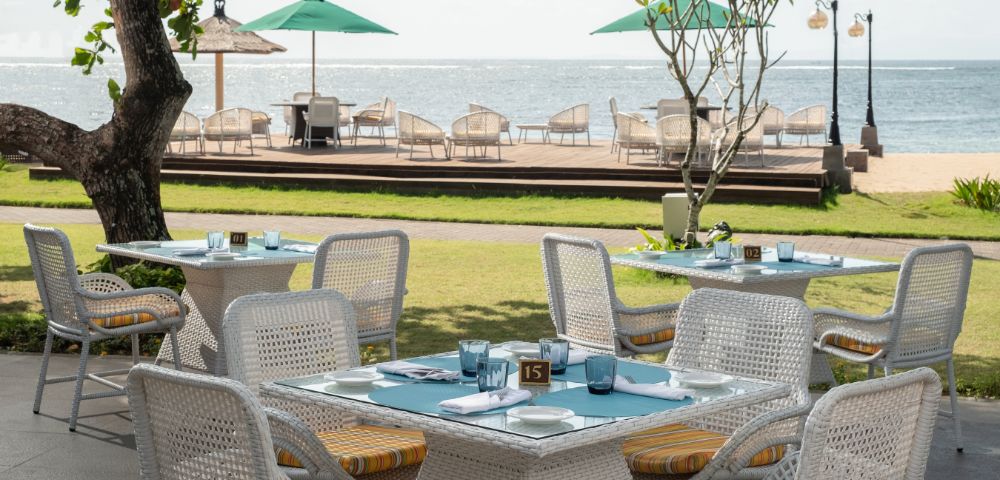 Outdoor dining area with wicker tables and chairs facing the ocean. Green umbrellas and deck chairs line the wooden deck. A tranquil, sunny atmosphere.
