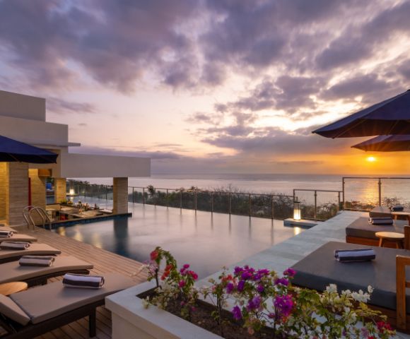 Rooftop infinity pool at sunset, with lounge chairs and umbrellas on a wooden deck. Vibrant flowers in the foreground and the ocean in the distance.