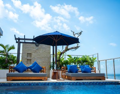 Rooftop pool area with two wooden lounge chairs, blue cushions, and blue umbrella under a clear sky. Tropical plants and ocean view in the background.