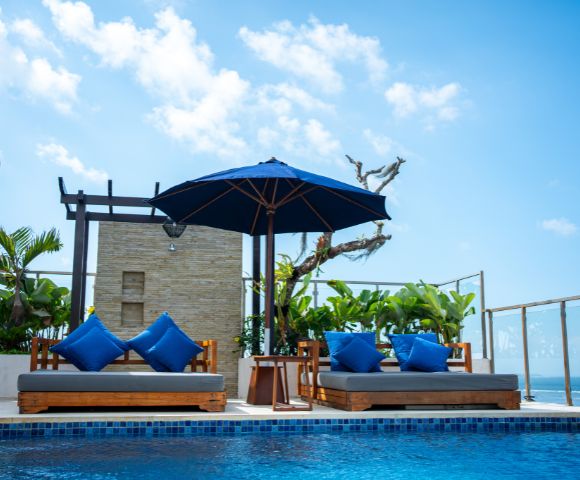 Rooftop pool area with two wooden lounge chairs, blue cushions, and blue umbrella under a clear sky. Tropical plants and ocean view in the background.