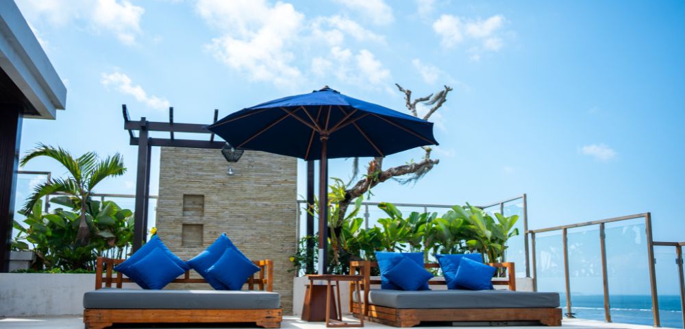Rooftop pool area with two wooden lounge chairs, blue cushions, and blue umbrella under a clear sky. Tropical plants and ocean view in the background.