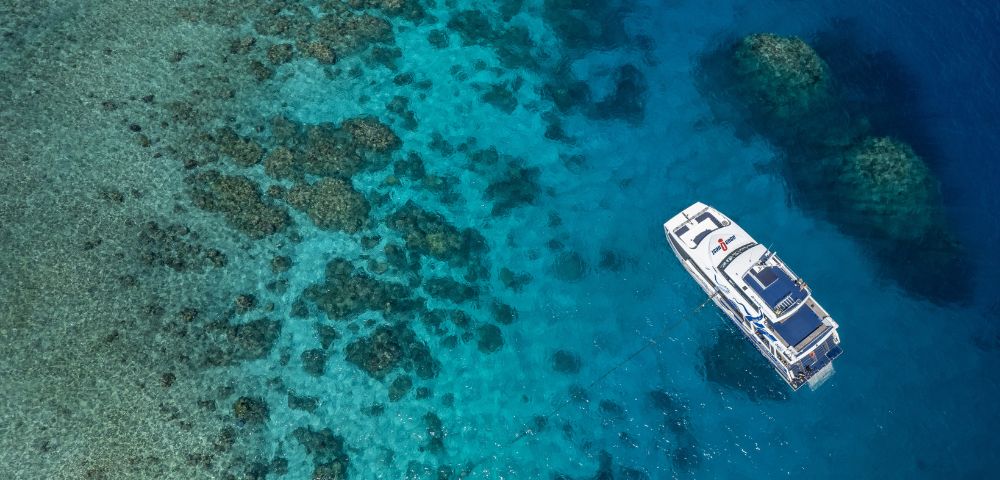 Aerial view of a white boat floating over vibrant, turquoise waters near coral reefs. The tranquil scene conveys serenity and the beauty of marine life.