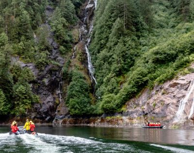 Two people on jet skis ride across a serene lake, bordered by lush green cliffs and a cascading waterfall, creating a sense of adventure and tranquility.