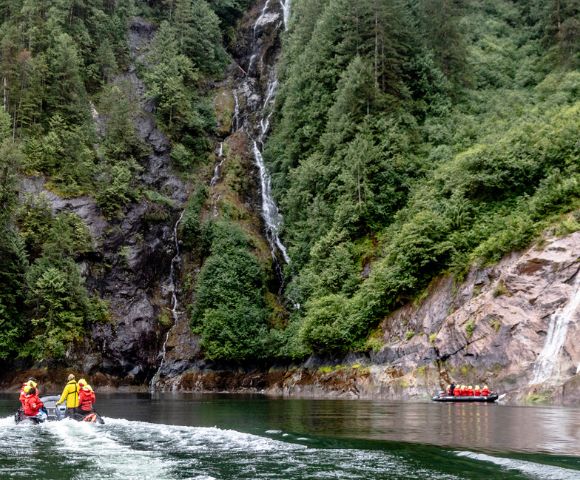 Two people on jet skis ride across a serene lake, bordered by lush green cliffs and a cascading waterfall, creating a sense of adventure and tranquility.