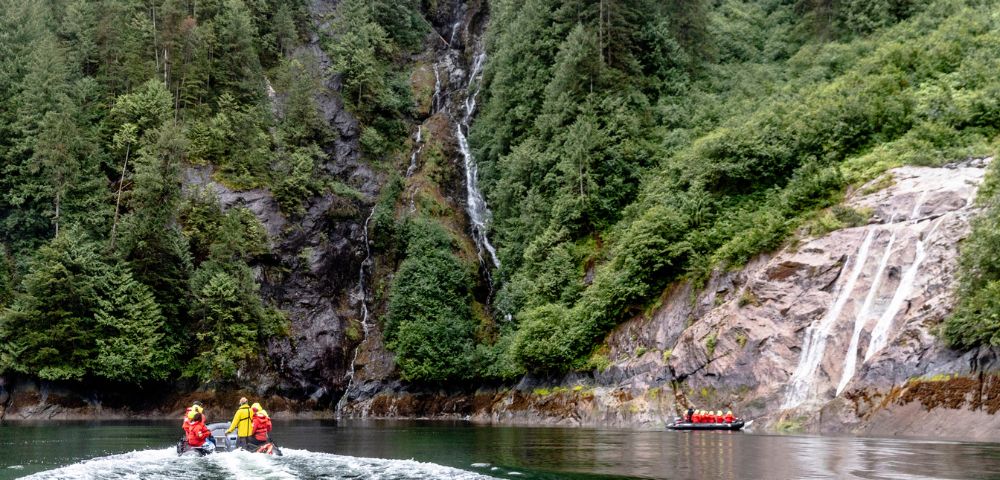Two people on jet skis ride across a serene lake, bordered by lush green cliffs and a cascading waterfall, creating a sense of adventure and tranquility.