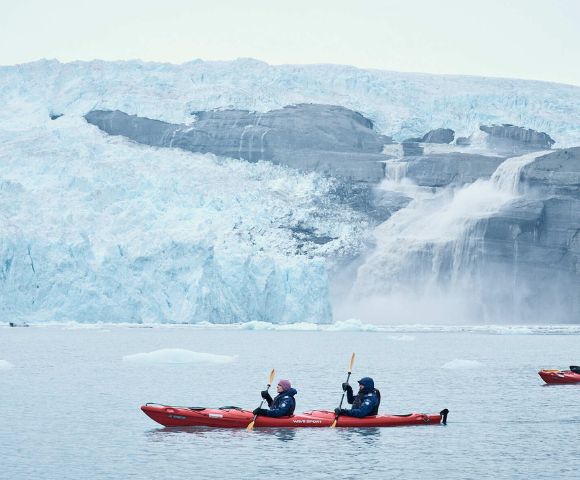 Two people in red kayaks paddle near a massive, jagged ice glacier with cascading water in a serene, cold setting, conveying adventure and tranquility.