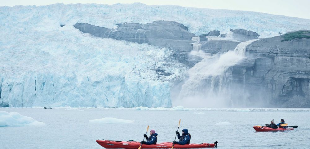 Two people in red kayaks paddle near a massive, jagged ice glacier with cascading water in a serene, cold setting, conveying adventure and tranquility.