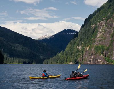 Two kayakers paddle on a tranquil lake surrounded by lush green mountains under a partly cloudy sky, evoking a sense of adventure and serenity.