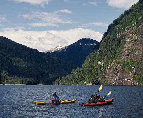 Two kayakers paddle on a tranquil lake surrounded by lush green mountains under a partly cloudy sky, evoking a sense of adventure and serenity.