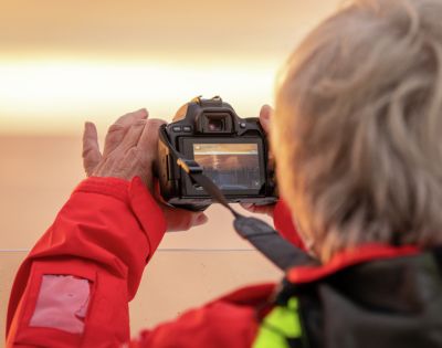 A person in a bright red jacket captures a photo using a digital camera. The camera screen displays a subtle scene, lit by warm, soft light, creating a focused and calm mood.
