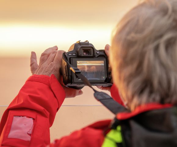 A person in a bright red jacket captures a photo using a digital camera. The camera screen displays a subtle scene, lit by warm, soft light, creating a focused and calm mood.