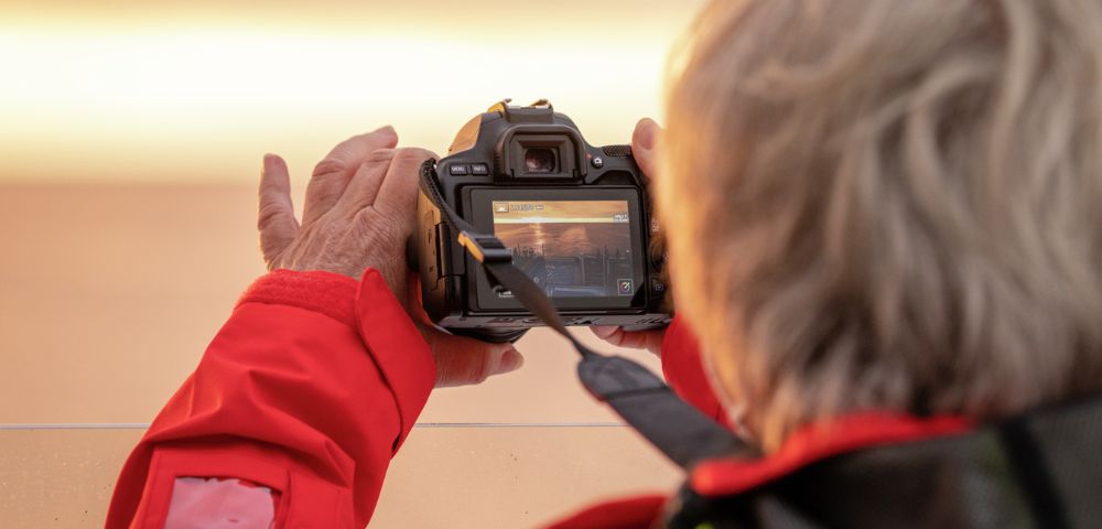 A person in a bright red jacket captures a photo using a digital camera. The camera screen displays a subtle scene, lit by warm, soft light, creating a focused and calm mood.