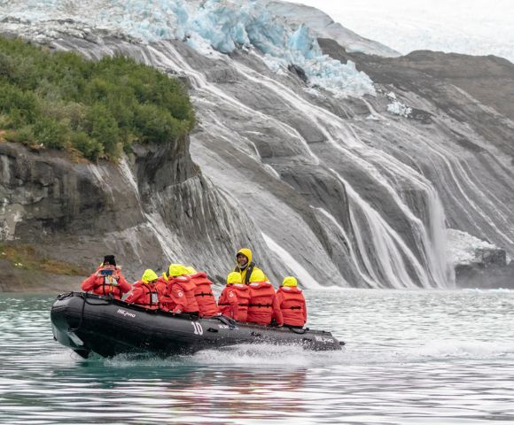 A group in red life jackets and yellow helmets are in a black inflatable boat on a turquoise lake near a rocky glacier with cascading water.