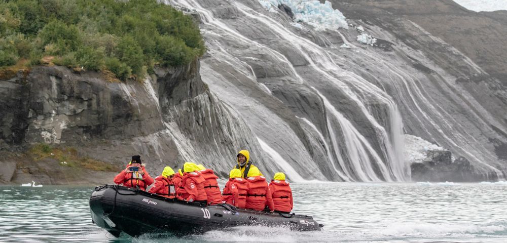 A group in red life jackets and yellow helmets are in a black inflatable boat on a turquoise lake near a rocky glacier with cascading water.