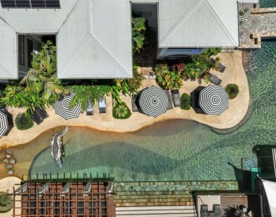 Overhead view of a tropical pool area with curvy paths, striped umbrellas, sun loungers, lush greenery, and a crocodile statue, creating a serene resort vibe.