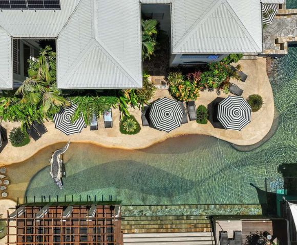 Overhead view of a tropical pool area with curvy paths, striped umbrellas, sun loungers, lush greenery, and a crocodile statue, creating a serene resort vibe.