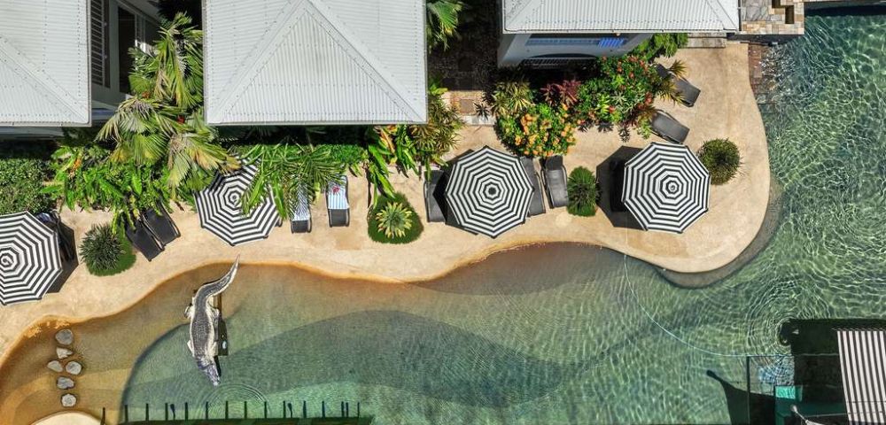 Overhead view of a tropical pool area with curvy paths, striped umbrellas, sun loungers, lush greenery, and a crocodile statue, creating a serene resort vibe.