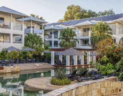 Tropical resort featuring white buildings with balconies, surrounded by lush greenery. A serene pool area with lounge chairs and umbrellas in the foreground.