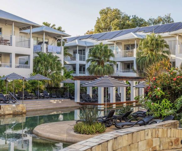 Tropical resort featuring white buildings with balconies, surrounded by lush greenery. A serene pool area with lounge chairs and umbrellas in the foreground.