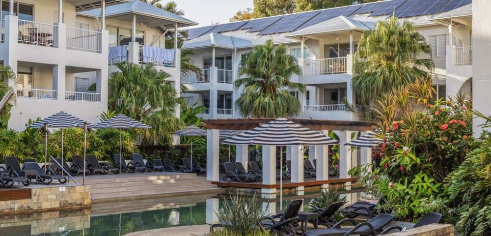 Tropical resort featuring white buildings with balconies, surrounded by lush greenery. A serene pool area with lounge chairs and umbrellas in the foreground.