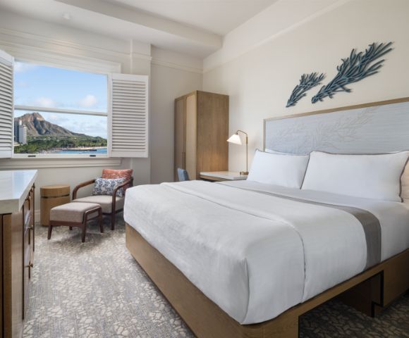 Bright hotel room with a large bed, modern furnishings, and a beach-themed decor. An open window reveals a scenic mountain view, offering a tranquil vibe.
