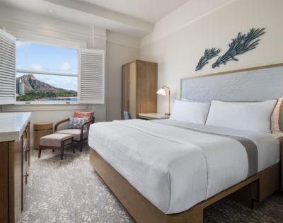 Bright hotel room with a large bed, modern furnishings, and a beach-themed decor. An open window reveals a scenic mountain view, offering a tranquil vibe.
